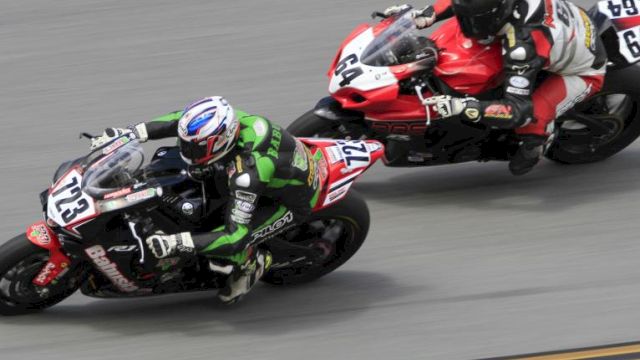 Two motorcyclists are racing on a track, one in green and black gear, the other in red and white, leaning into a curve.