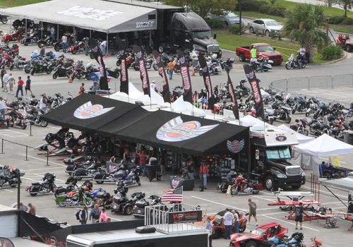 The image shows a busy motorcycle event with various bikes, tents, flags, and people gathered in an outdoor setting.