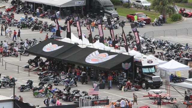 The image shows a busy motorcycle event with various bikes, tents, flags, and people gathered in an outdoor setting.