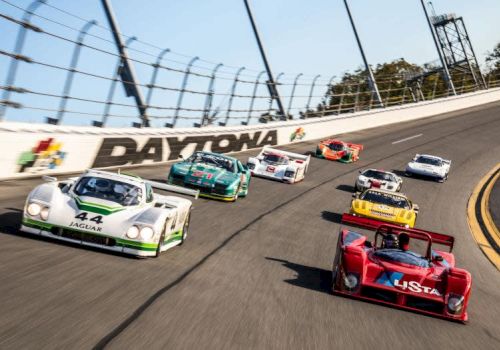 The image shows a group of race cars on a track with "Daytona" written on a wall, racing around a curve in a competitive event.