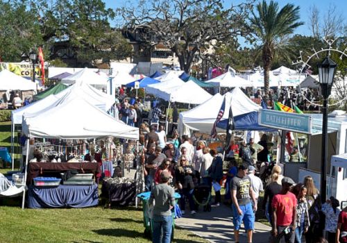The image shows a crowded outdoor market with numerous white tents and people walking around on a sunny day.