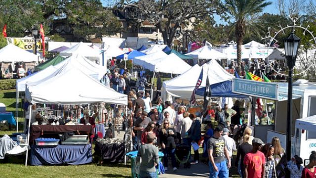 The image shows a crowded outdoor market with numerous white tents and people walking around on a sunny day.