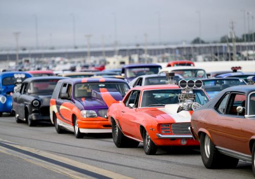 A lineup of colorful, customized cars is parked on a race track, featuring various classic and modern vehicles in vibrant hues.