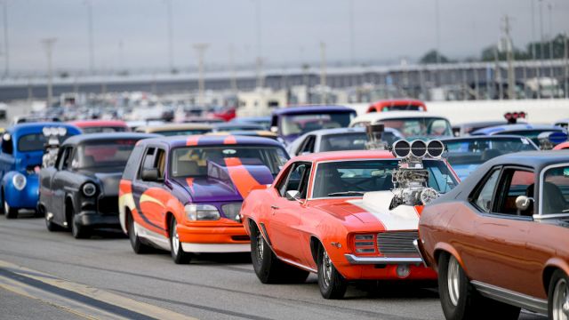 A lineup of colorful, customized cars is parked on a race track, featuring various classic and modern vehicles in vibrant hues.