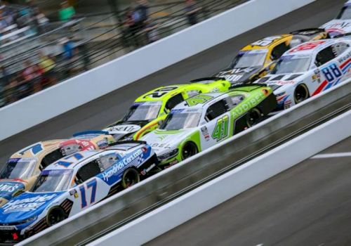 A high-speed stock-car race shows multiple colorful NASCAR cars speeding on a paved track under motion blur.