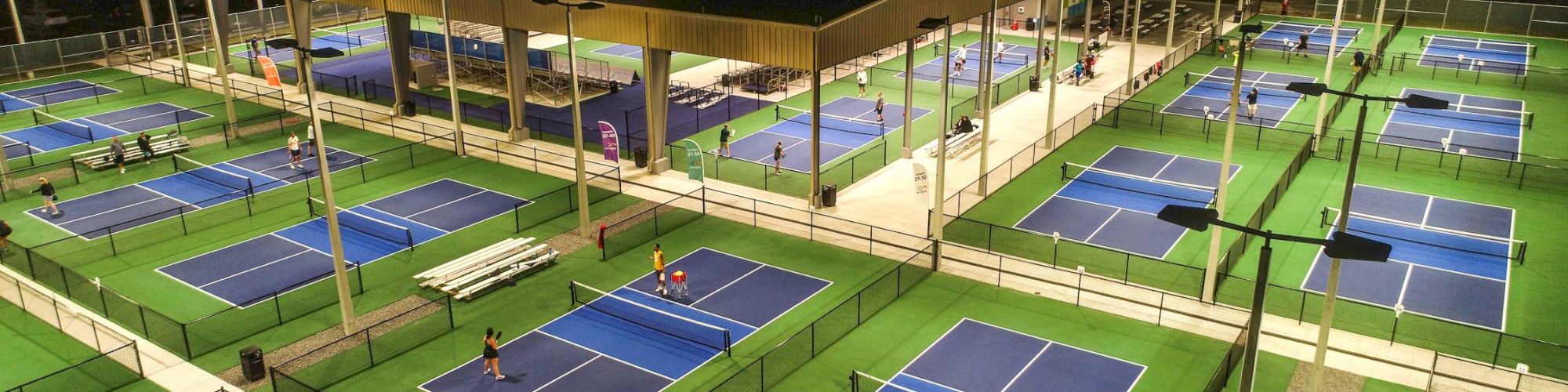 An illuminated court complex at night with multiple pickleball courts and people playing, surrounded by benches and a covered area.