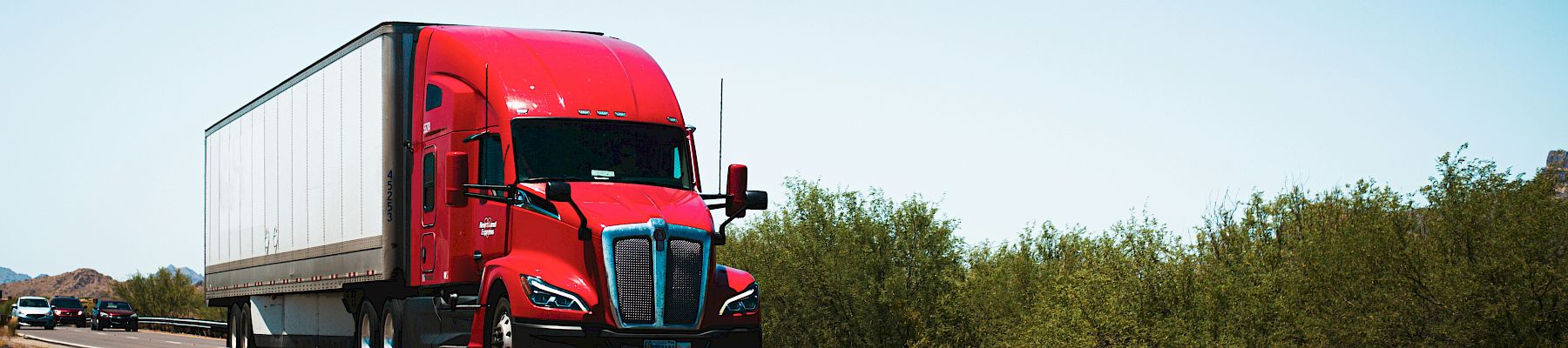 A red semi-truck driving on a highway, with vegetation and a clear blue sky in the background, captured during daytime.