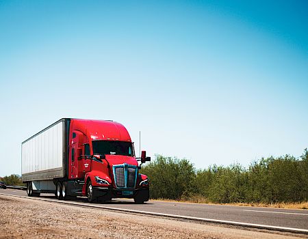 A red semi-truck driving on a highway, with vegetation and a clear blue sky in the background, captured during daytime.