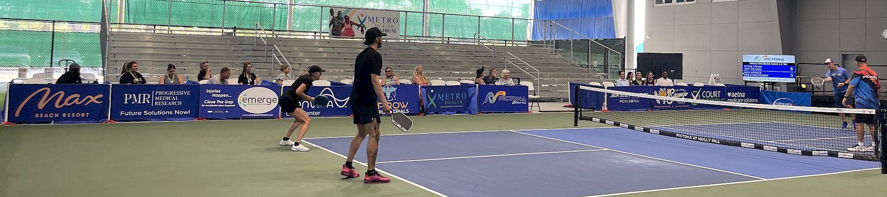 An indoor tennis match is taking place with spectators in the stands. The court is surrounded by banners and a high ceiling.