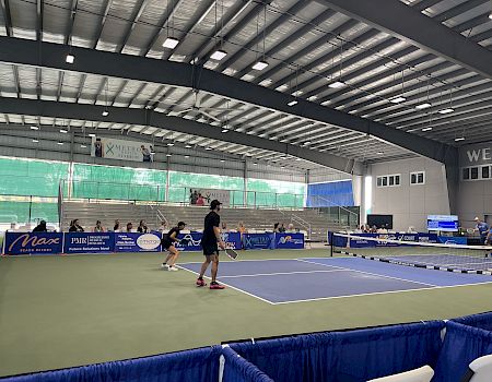 An indoor tennis match is taking place with spectators in the stands. The court is surrounded by banners and a high ceiling.