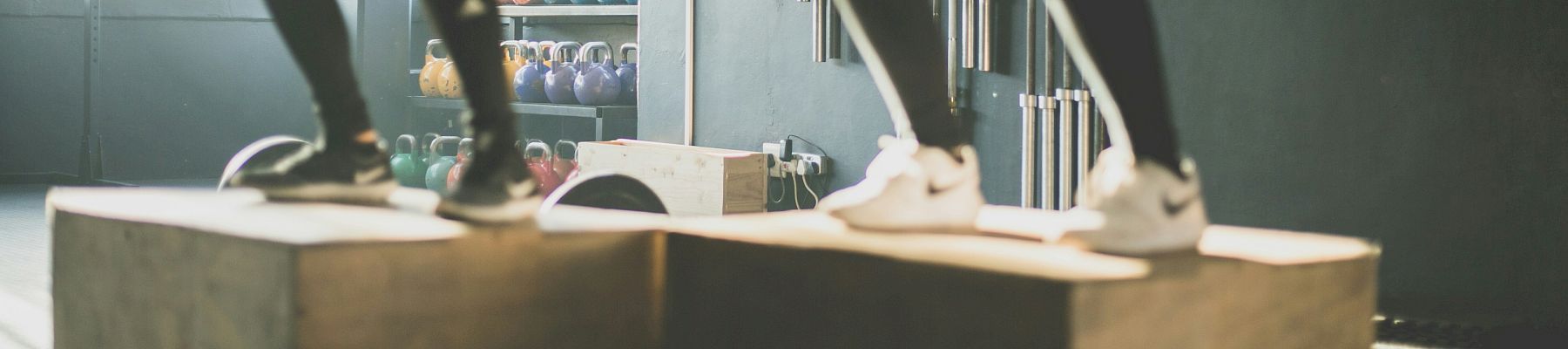 Two people are performing box jumps in a gym with "Raise the Bar" on the wall in the background.