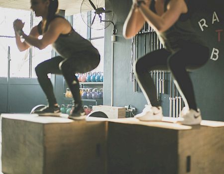 Two people are performing box jumps in a gym with "Raise the Bar" on the wall in the background.