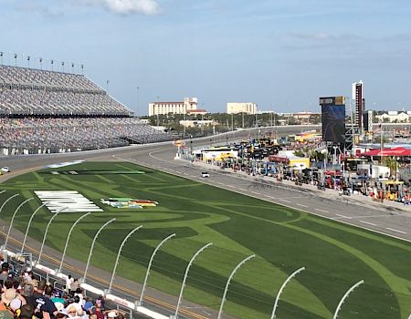 A car race is underway at a large stadium with spectators in the stands and a clear blue sky.