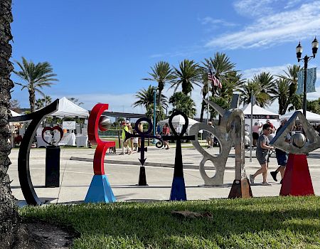 Sculptures are set up in a park with people walking, tents, palm trees, and a blue sky in the background.