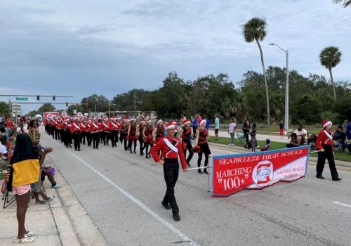 A marching band in red uniforms parades down a street, with onlookers on the side, and a banner leading the group.