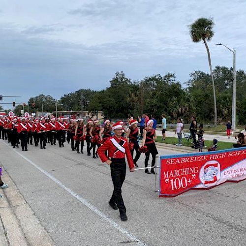 A marching band in red uniforms parades down a street, with onlookers on the side, and a banner leading the group.