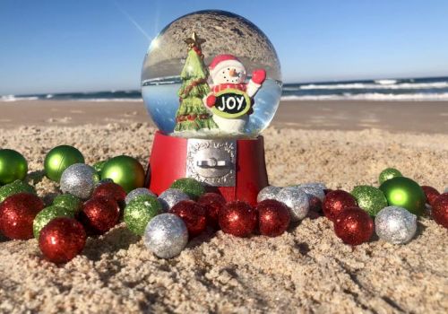 A snow globe with a Santa and tree sits on a beach surrounded by colorful ornaments, with the ocean in the background.