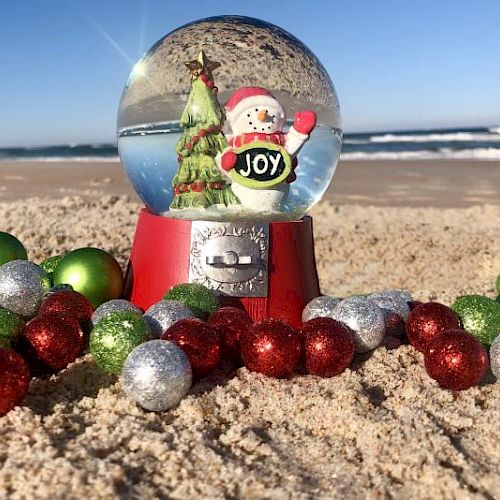 A snow globe with a Santa and tree sits on a beach surrounded by colorful ornaments, with the ocean in the background.