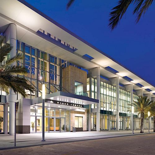 A modern building with large windows is illuminated at night, surrounded by palm trees, under a clear evening sky.