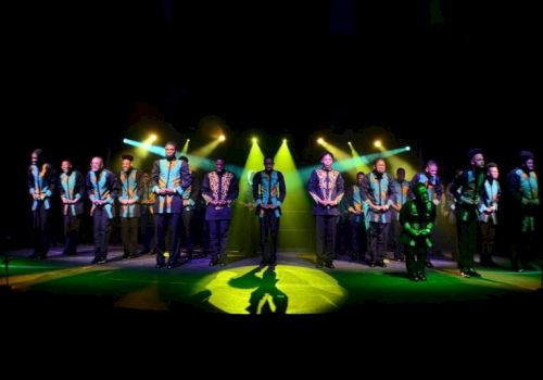 A group of musicians/performers in patterned shirts stand together on a concert stage under bright green/yellow spotlights.