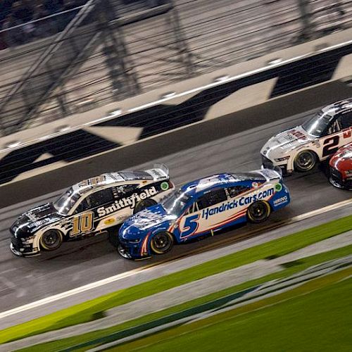 Several race cars are competing on a track, closely packed together, with a blurred background indicating high speed action.