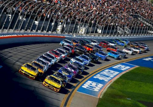 A packed NASCAR race track shows many colorful stock cars speeding past grandstands and a &ldquo;Coca-Cola&rdquo; branded curve.
