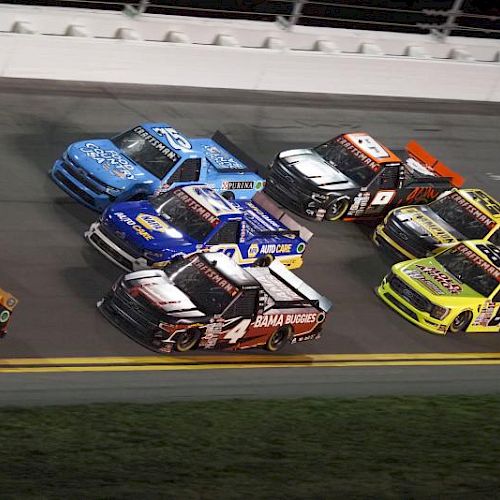 The image shows a group of racing trucks on a track, competing closely in a NASCAR event at night, with various colors and logos.
