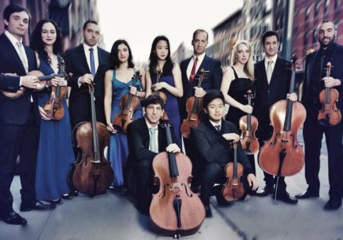 A group of musicians stands in formal attire on a city street, holding string instruments like violins, violas, and cellos.