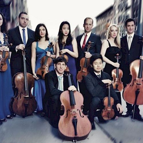 A group of musicians stands in formal attire on a city street, holding string instruments like violins, violas, and cellos.