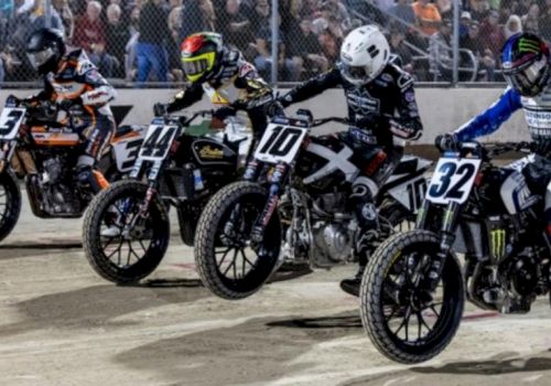 Motorcyclists lined up for a race on dirt track. Each bike has numbers, riders with helmets. Spectators watching from behind the fence.