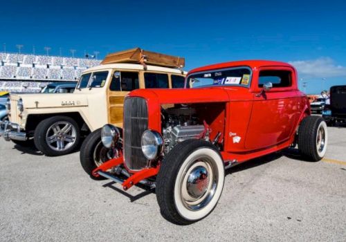 A bright red vintage hot rod and a classic cream car with a wooden roof are parked on a sunny day, showcasing their retro style.