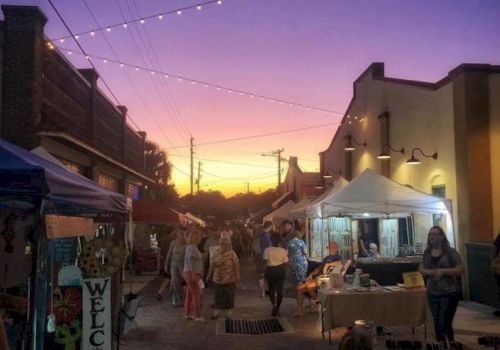 A sunset street market with string lights, vendor tents, and shoppers walking along the road.