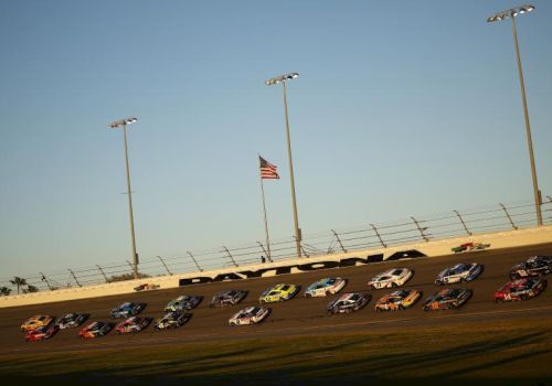 A NASCAR-style race track with many stock cars parked in a row under bright stadium lights and an American flag flying.