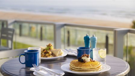 A table with pancakes, a breakfast wrap, coffee mugs, and water sits near a beach view for a relaxing meal experience.
