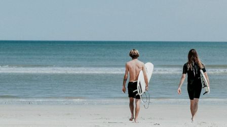 Two people walk toward the ocean, each carrying a surfboard, on a calm, sandy beach under a clear sky.
