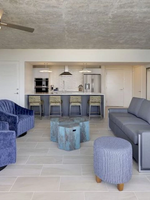 The image shows a modern living room with purple chairs, a sofa, a coffee table, kitchen island with stools, ceiling fan, and tiled floor.