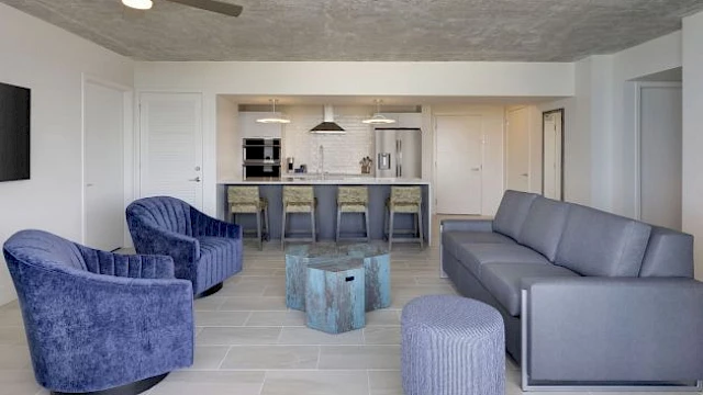 The image shows a modern living room with purple chairs, a sofa, a coffee table, kitchen island with stools, ceiling fan, and tiled floor.
