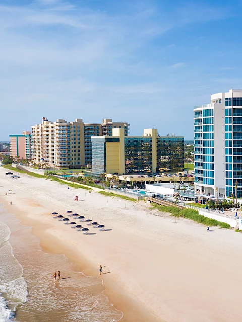 A beach scene with people, a sandy shore, ocean waves, and several high-rise buildings lining the coastline under a clear blue sky.