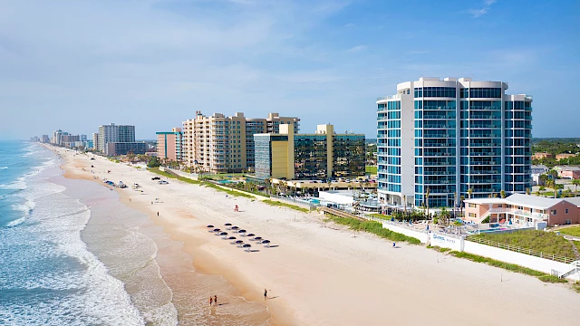 A beach scene with people, a sandy shore, ocean waves, and several high-rise buildings lining the coastline under a clear blue sky.