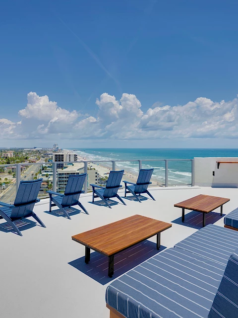 A rooftop terrace with lounge chairs and tables, offering a panoramic view of the coastline and ocean under a partly cloudy sky.