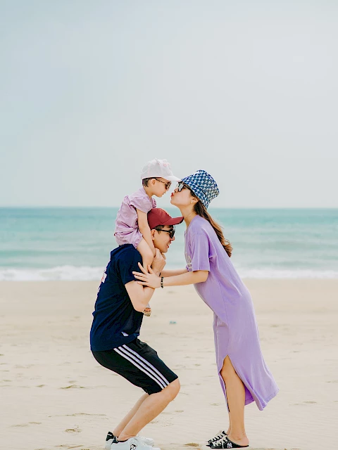 A family at the beach: a person piggybacks a child while another person kisses the child's head, all wearing casual attire and hats.