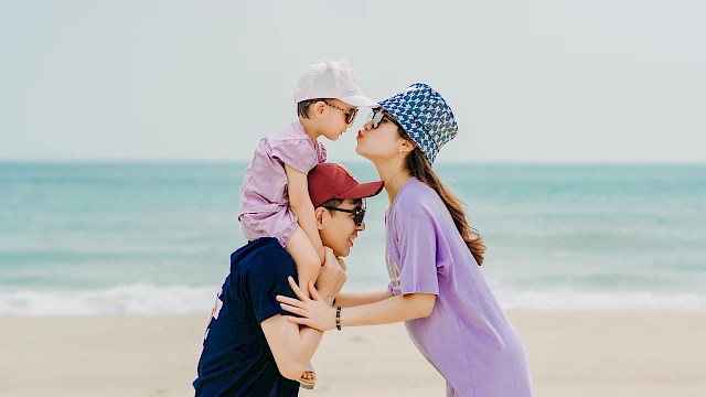 A family at the beach: a person piggybacks a child while another person kisses the child's head, all wearing casual attire and hats.