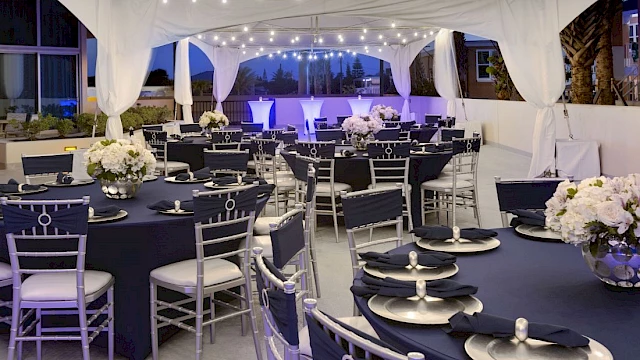 An elegant event setup under a white tent with round tables, navy decor, white flowers, and string lights creating a sophisticated ambiance.