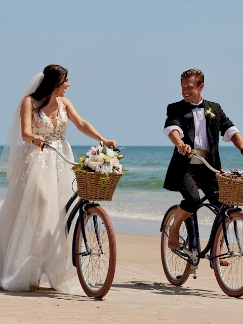 A bride and groom are joyfully riding bicycles on a sandy beach, with the ocean in the background, each having a basket on the bike.