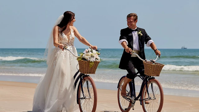 A bride and groom are joyfully riding bicycles on a sandy beach, with the ocean in the background, each having a basket on the bike.