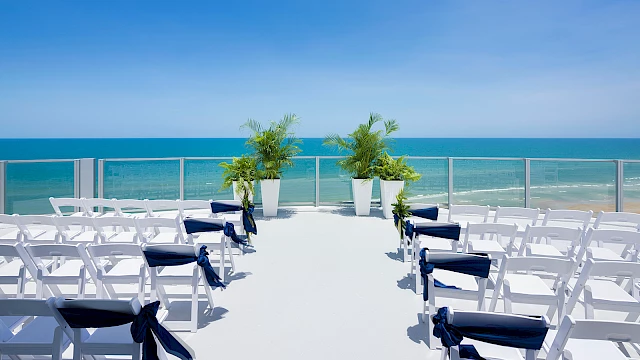 Outdoor wedding setup with white chairs and navy ribbons facing the ocean, under a clear sky, with two tall potted plants at the front.