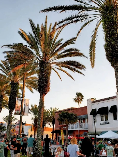 A lively street scene with palm trees, people walking, and outdoor activities under a clear sky, with buildings lining the street.