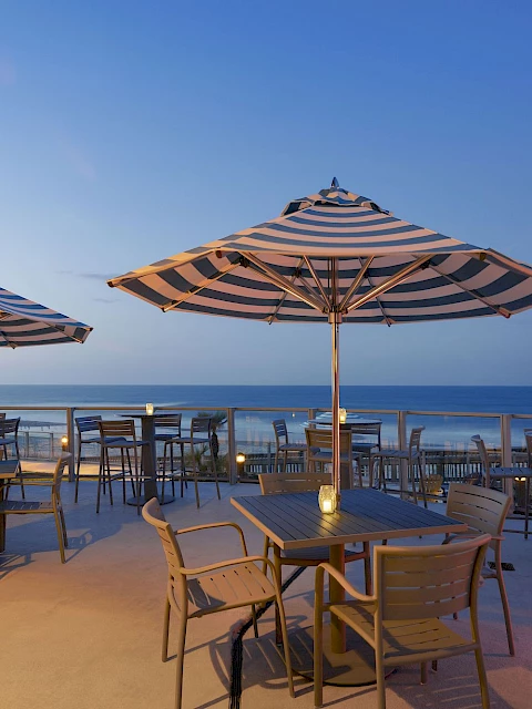 Outdoor patio with tables, striped umbrellas, and chairs overlooking the ocean at sunset under a clear sky, creating a serene atmosphere.