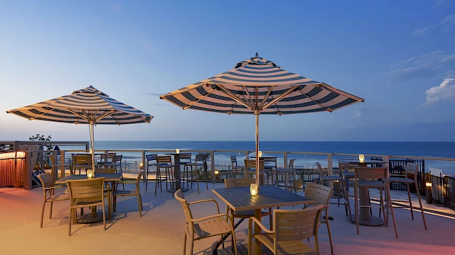 Outdoor patio with tables, striped umbrellas, and chairs overlooking the ocean at sunset under a clear sky, creating a serene atmosphere.