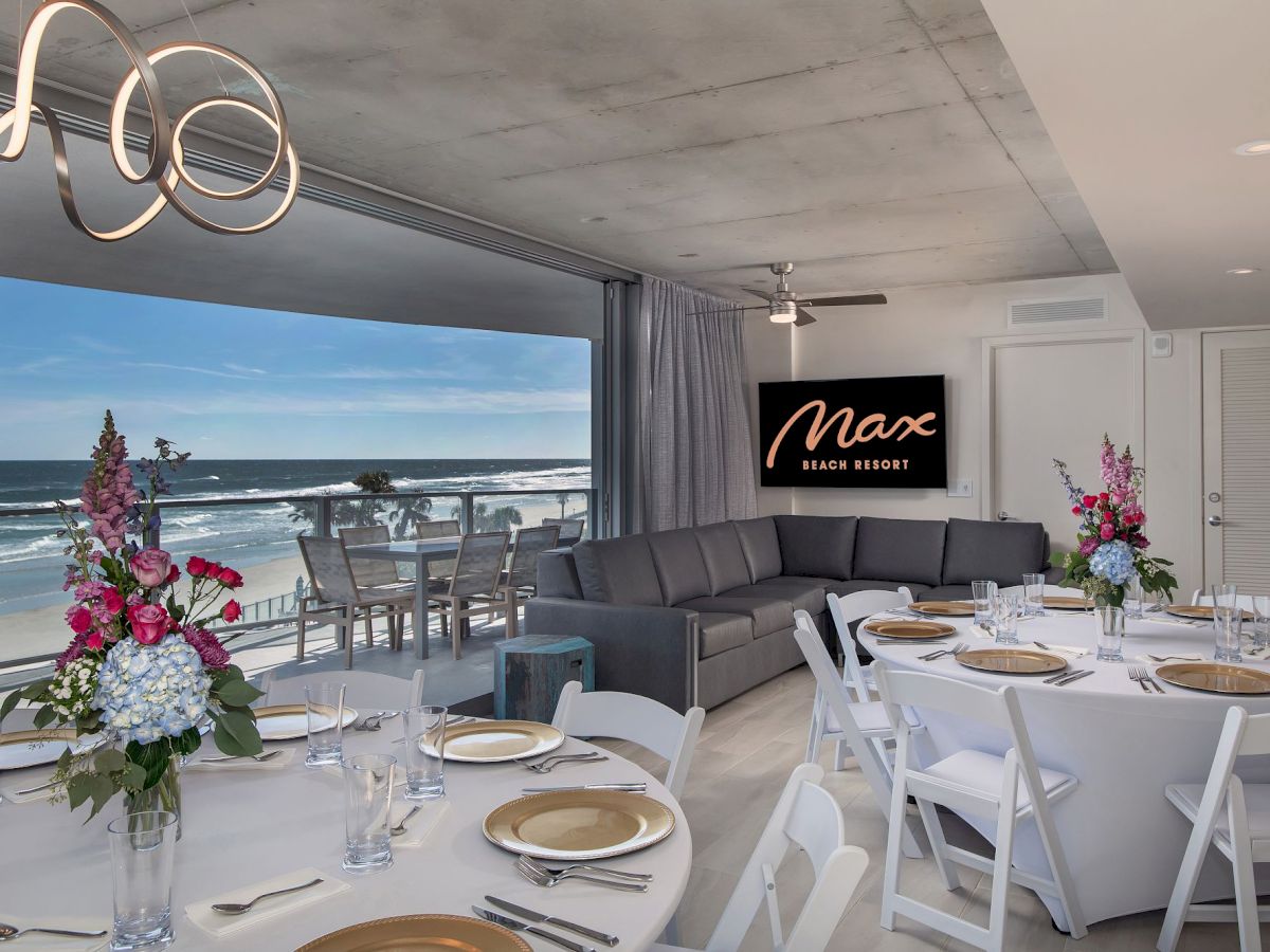 The image shows a seaside event space with elegant table settings and floral arrangements on round tables, a couch, and a "Max" sign on the wall.